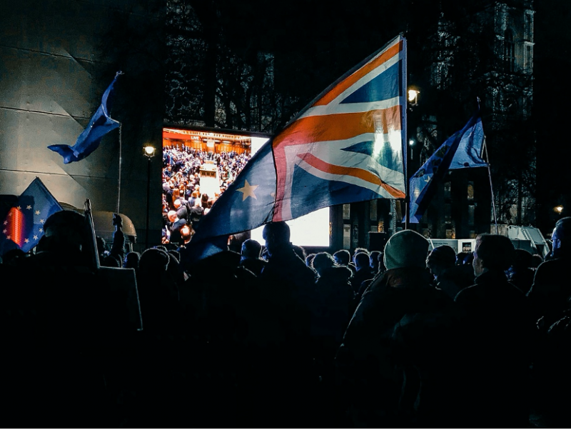 A crowd fo people stand in front of a large TV screen waiving the British flag